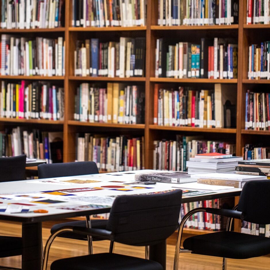 Priekšplānā ir galds ar krēsliem un tālākajā plānā ir liels grāmatu plaukts